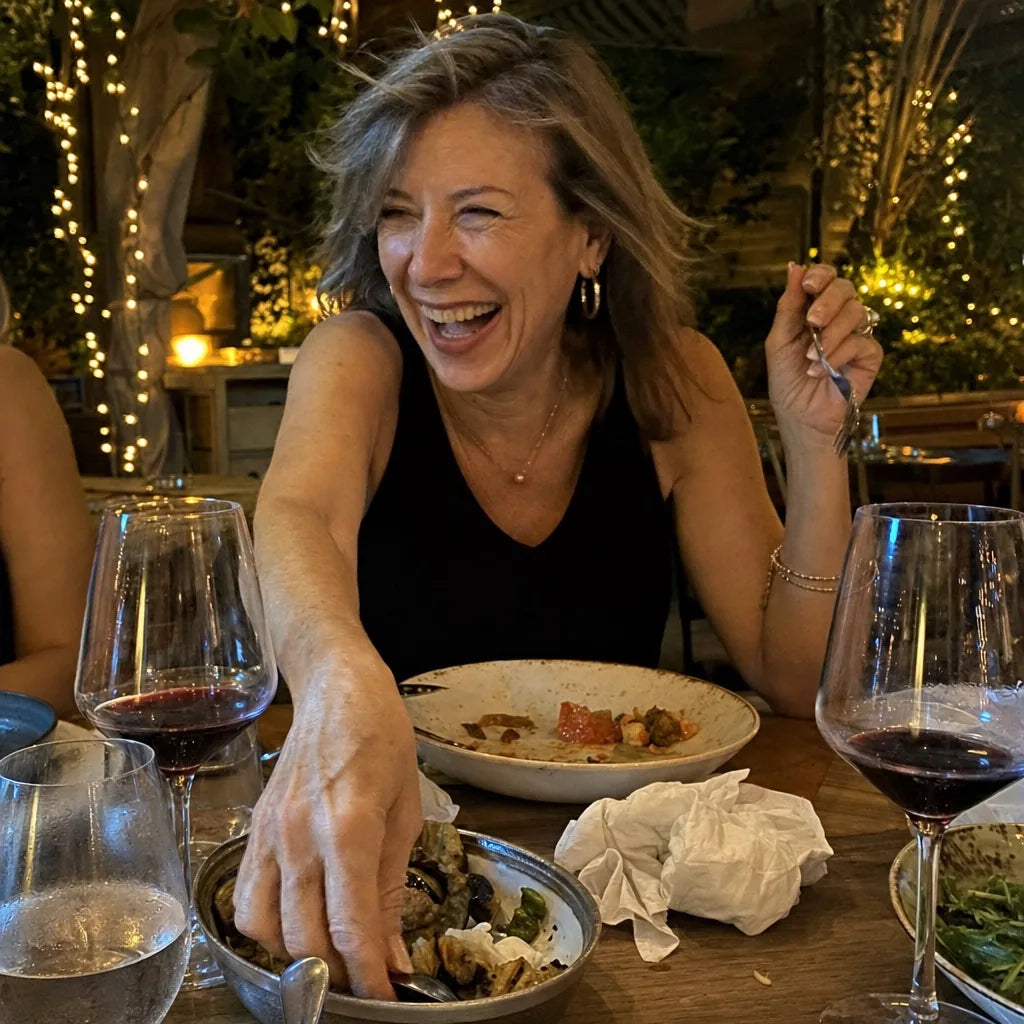 A woman laughing at dinner with friends, reaching for a shared appetizer
